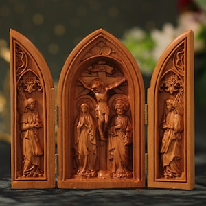 Puede incluir: Un tríptico de madera con una imagen tallada de Jesús en la cruz, flanqueado por María y Juan. El tríptico está cerrado, mostrando las figuras talladas de ángeles en los paneles laterales.