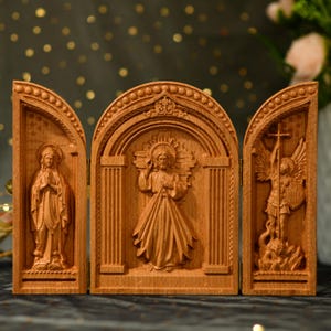Op de afbeelding: Een houten triptiek met drie gegraveerde panelen die religieuze figuren voorstellen. Het middelste paneel toont Jezus Christus met zijn hart bloot, het linker paneel toont de Maagd Maria en het rechter paneel toont een engel met een zwaard.