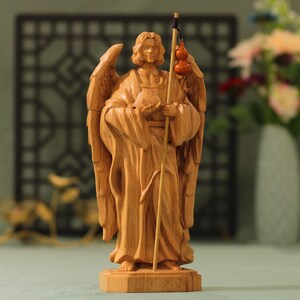 May include: A wooden sculpture of an angel with wings, holding a staff and a gourd. The angel is standing on a wooden base.
