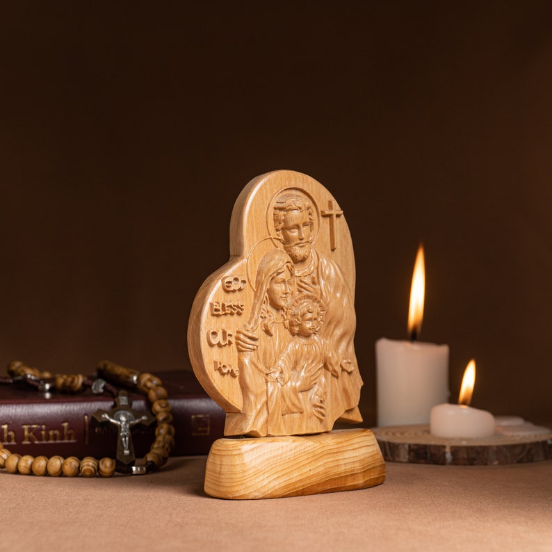 Op de afbeelding: Een houten beeldhouwwerk van de Heilige Familie, Jezus, Maria en Jozef, in de vorm van een hart. Het beeldhouwwerk staat op een houten voet en heeft de woorden "God zegen ons huis" erop gegraveerd.