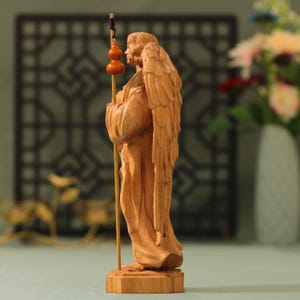 May include: A wooden sculpture of an angel with wings, holding a staff with a gourd on top. The angel is standing on a wooden base.