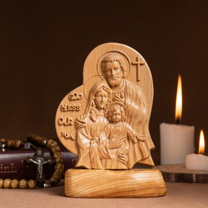 Op de afbeelding: Een houten beeldhouwwerk van de Heilige Familie, Jezus, Maria en Jozef, in de vorm van een hart. Het beeldhouwwerk staat op een houten voet en heeft de woorden "God zegen ons huis" erop gegraveerd.