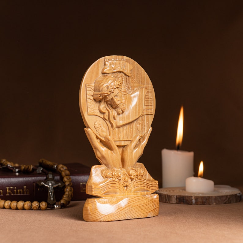 May include: A wooden sculpture of Jesus Christ on the cross, with a crown of thorns, surrounded by hands holding a heart. The sculpture is on a wooden base with a book carved into it. The sculpture is in front of two lit candles.