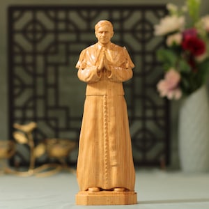 May include: A wooden statue of a man in religious attire, with hands clasped in prayer. The figure is carved from light-colored wood, showcasing the grain. The statue stands on a rectangular base, with a detailed robe and cape.