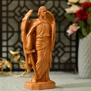 May include: A wooden sculpture of an angel with wings, holding a sword and a shield. The angel is carved in a standing position with a serious expression on its face.