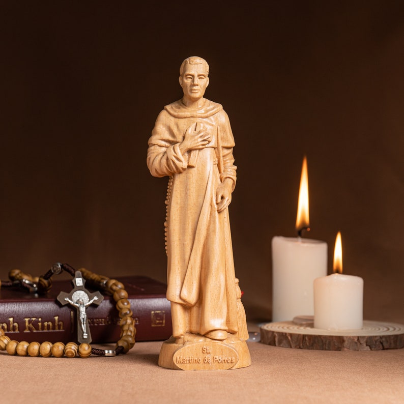 May include: A wooden statue of Saint Martin de Porres, a Catholic saint, standing with his hands clasped in prayer. The statue is on a wooden base with the inscription "St. Martino de Porres". The statue is surrounded by two lit candles and a rosary.
