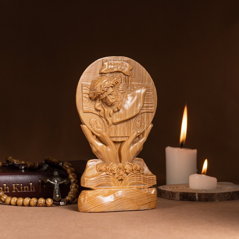 May include: A wooden sculpture of Jesus Christ on the cross, with a crown of thorns, held in the hands of a person. The sculpture is on a wooden base with a book carved into it. The sculpture is in front of two white candles.