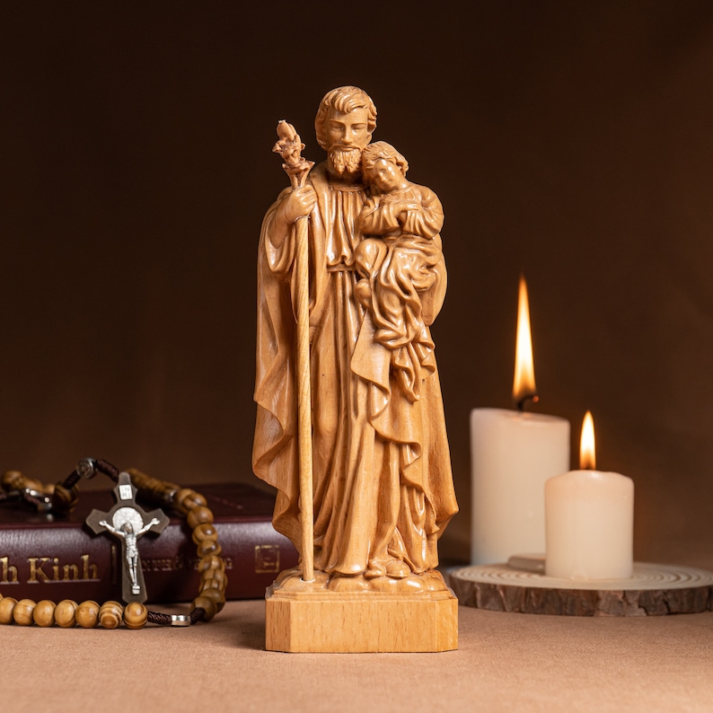 May include: A wooden statue of Saint Joseph holding a child. The statue is light brown and has a simple, traditional design. The statue is standing on a wooden base.