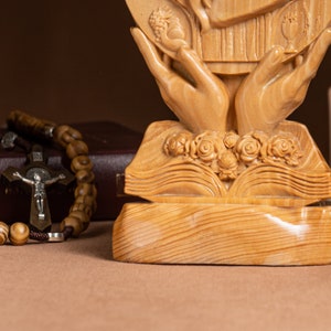 May include: A wooden sculpture of two hands holding a book with a carved image of a heart and flowers. The sculpture is on a wooden base.