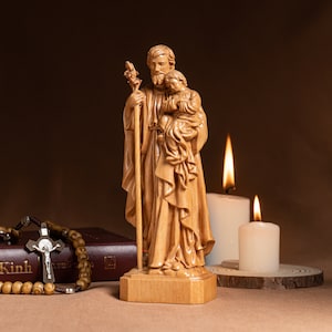 May include: A wooden statue of Saint Joseph holding a child. The statue is standing on a wooden base and is surrounded by candles.