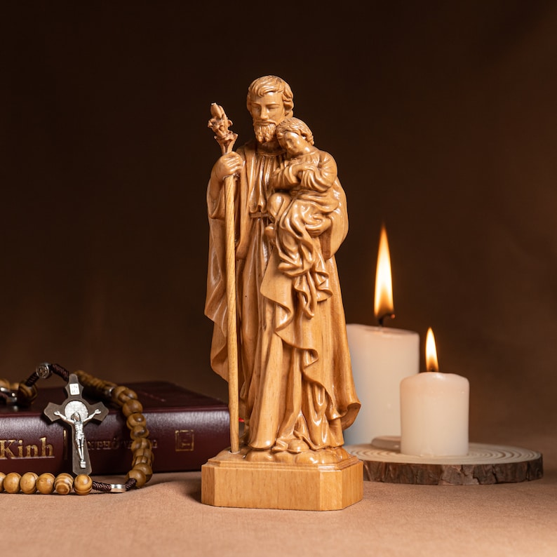 May include: A wooden statue of Saint Joseph holding a child. The statue is standing on a wooden base and is surrounded by candles.