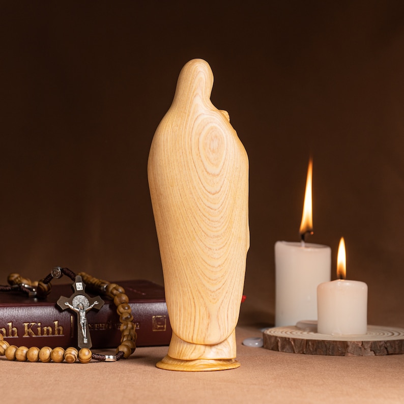 May include: A wooden statue of the Virgin Mary with a child in her arms. The statue is light brown and has a smooth, polished finish. The statue is standing on a wooden base. Two white candles are burning in the background.