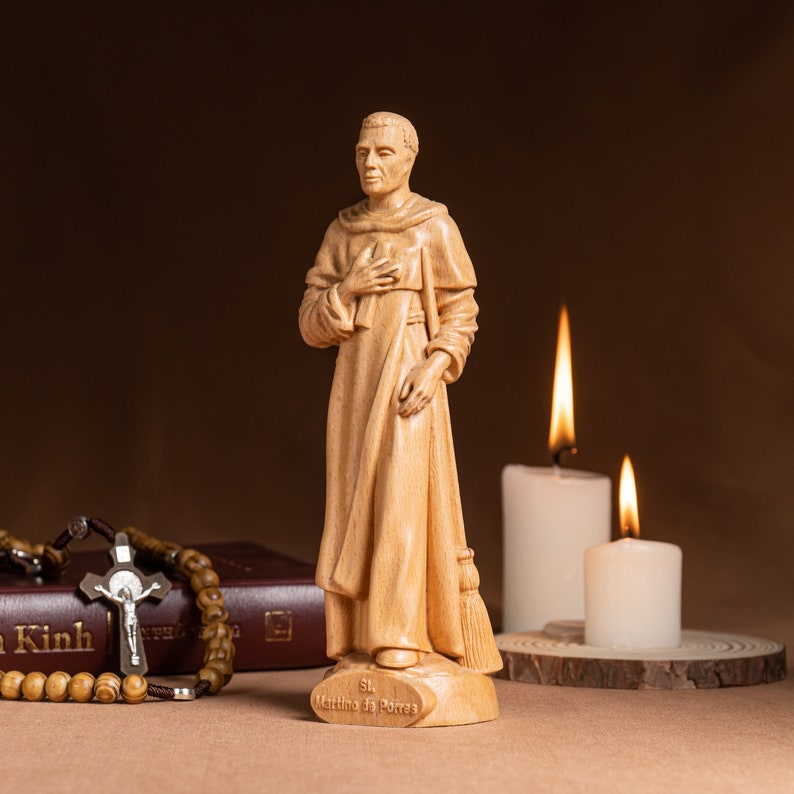 May include: A wooden statue of Saint Martin de Porres, a Catholic saint, standing with his hands clasped in prayer. The statue is on a wooden base with the inscription "St. Martin de Porres". The statue is in front of two white candles.