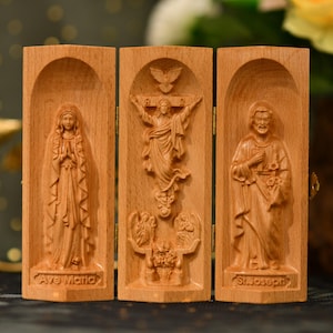 Może przedstawiać: Drewniany tryptyk z rzeźbionymi postaciami religijnymi. Lewy panel głosi "Ave Maria" i przedstawia postać ze złożonymi dłońmi. Środkowy panel przedstawia postać na krzyżu. Prawy panel głosi "St. Joseph".
