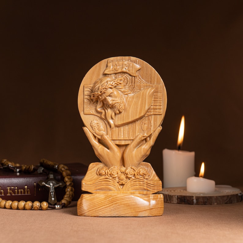 May include: A wooden sculpture of Jesus Christ on the cross, with a crown of thorns, surrounded by two hands holding a chalice and a host. The sculpture is on a wooden base with a book and flowers carved into it. The sculpture is in front of a brown background.