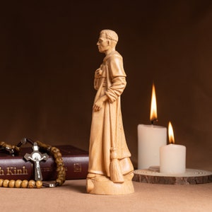 May include: A wooden figurine of a man in a robe, holding a broom. The figurine is standing on a wooden base. There are two candles in the background, one lit and one unlit.