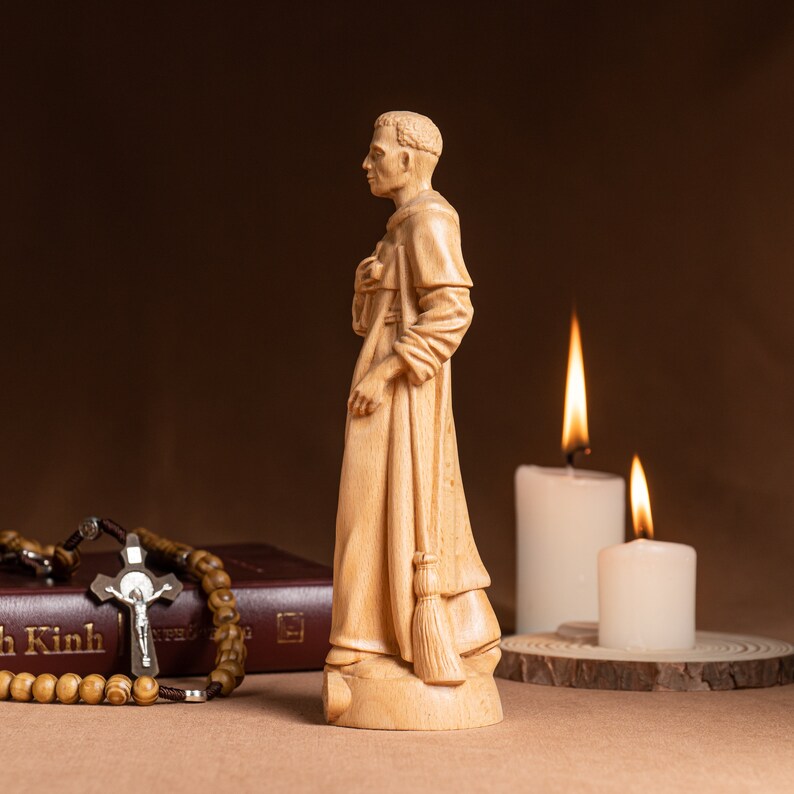 May include: A wooden figurine of a man in a robe, holding a broom. The figurine is standing on a wooden base. There are two candles in the background, one lit and one unlit.