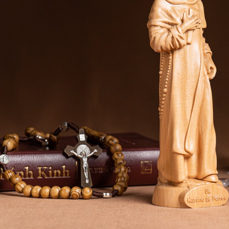 May include: A wooden figurine of a saint holding a rosary in their hand. The figurine is standing on a wooden base with the inscription "St. Martine de Porres". A wooden rosary with a crucifix is lying on a brown book with the text "kinh kinh" on the cover.