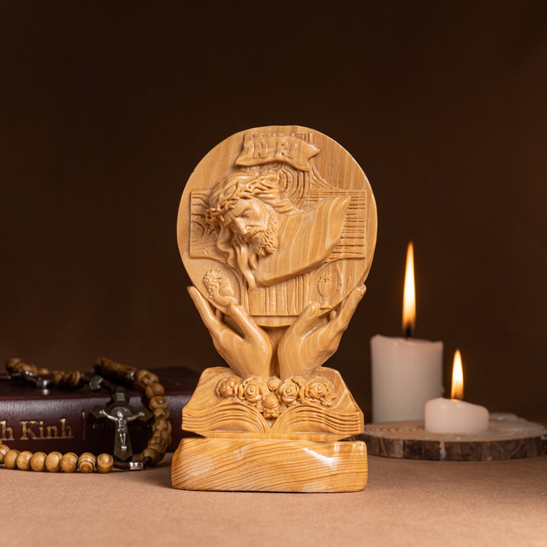 May include: A wooden sculpture of Jesus Christ on the cross, with a crown of thorns, surrounded by a circle of hands holding a chalice and a host. The sculpture is on a wooden base with a book and flowers.