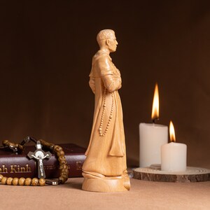 May include: A wooden statue of a person wearing a robe and holding a rosary. The statue is standing on a wooden base. There are two lit candles in the background.
