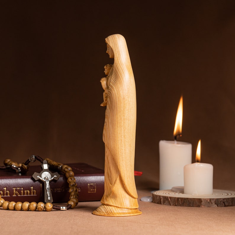May include: A wooden statue of the Virgin Mary holding a baby Jesus. The statue is light brown and has a detailed wood grain pattern. The statue is standing on a wooden base. Two white candles are in the background.