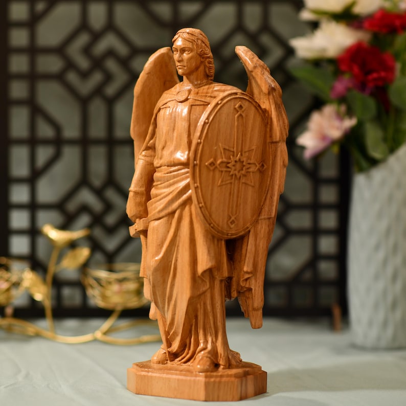 May include: A wooden sculpture of an angel with wings outstretched, holding a shield with a cross and star design. The angel is carved in a light brown color and stands on a wooden base.