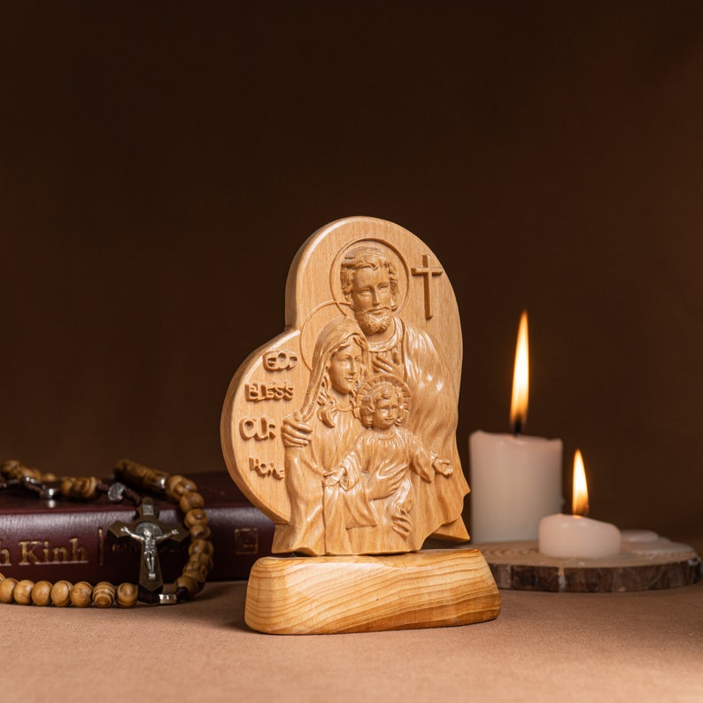 Op de afbeelding: Een houten beeldhouwwerk van de Heilige Familie, met Jezus, Maria en Jozef, in een hartvormig frame. Het beeldhouwwerk staat op een houten voet en is omringd door twee brandende kaarsen.