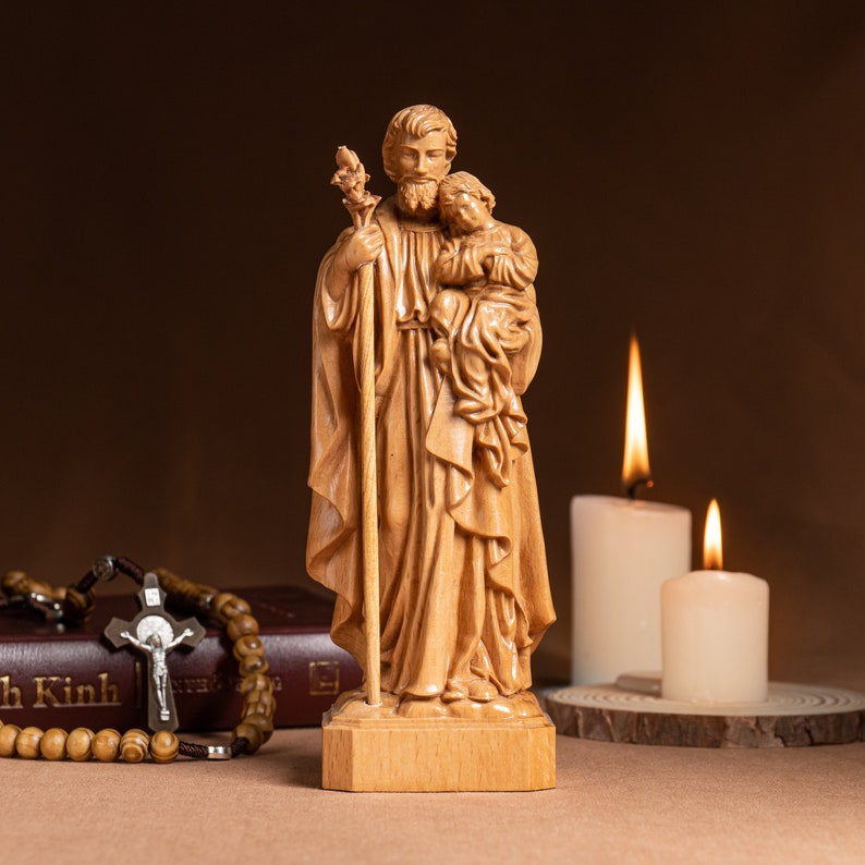 May include: A wooden statue of Saint Joseph holding a child. The statue is light brown and has a simple, traditional design. The statue is standing on a wooden base.