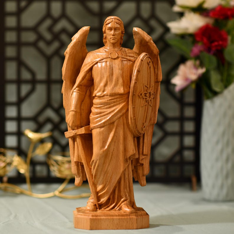 May include: A wooden sculpture of an angel with wings outstretched, holding a sword and a shield. The angel is carved in a realistic style and has a serene expression.