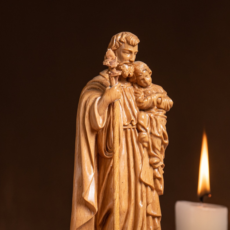 May include: A wooden statue of Saint Christopher carrying a child on his shoulder. The statue is carved in detail and has a warm brown color. A lit candle is in the background.