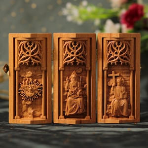 Könnte beinhalten: Ein dreiteiliges Holztriptychon mit drei Tafeln, die jeweils ein geschnitztes Bild einer religiösen Figur zeigen. Die mittlere Tafel zeigt eine Figur mit einer Krone und einem Stab, während die linke und rechte Tafel Figuren mit Kronen und Kreuzen zeigen. Die Tafeln sind mit aufwendigen Schnitzereien verziert.