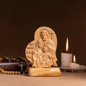 Op de afbeelding: Een houten beeldhouwwerk van de Heilige Familie, Jezus, Maria en Jozef, in de vorm van een hart. Het beeldhouwwerk staat op een houten voet en heeft de woorden "God zegen ons huis" in het hout gegraveerd. Het beeldhouwwerk staat voor twee witte kaarsen.