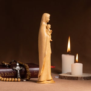 May include: A wooden statue of the Virgin Mary holding a baby Jesus. The statue is standing on a table with a book and rosary beads in front of it. Two white candles are lit in the background.