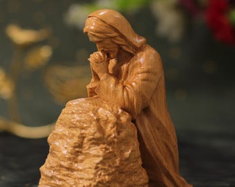 May include: A wooden sculpture of a person kneeling in prayer. The sculpture is carved in a realistic style and depicts the person with their head bowed and hands clasped in prayer. The sculpture is made of a light brown wood and has a natural finish.