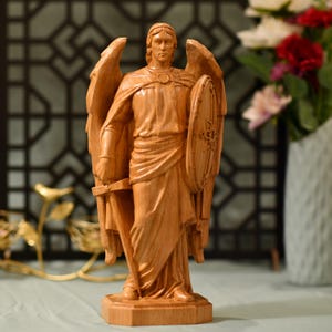 May include: A wooden statue of an angel with wings, holding a sword and a shield. The angel is carved in a standing position with a flowing robe and a serious expression.