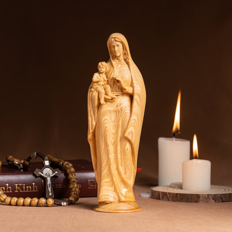 May include: A wooden statue of the Virgin Mary holding a baby Jesus. The statue is carved with intricate details and has a warm, natural wood finish. The statue is displayed on a table with a rosary and two candles.
