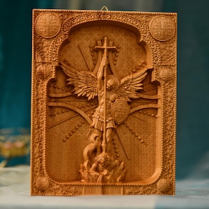 May include: A wooden carving of the Archangel Michael, a warrior angel with wings, holding a spear and shield. The carving is detailed and intricate, with a cross behind the angel and a dragon at the base.