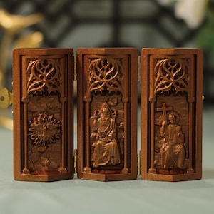 Könnte beinhalten: Ein dreiteiliges Holztriptychon mit drei geschnitzten Tafeln, die religiöse Figuren darstellen. Die mittlere Tafel zeigt eine auf einem Thron sitzende Figur, die linke Tafel zeigt eine Taube mit Lichtstrahlen und die rechte Tafel zeigt eine Figur, die ein Kreuz hält.