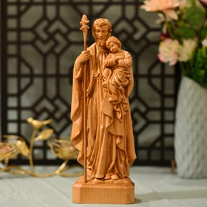 Può includere: Una scultura in legno di San Giuseppe che tiene in braccio un bambino Gesù. La scultura è scolpita in uno stile realistico ed è dipinta in un tono di legno naturale.