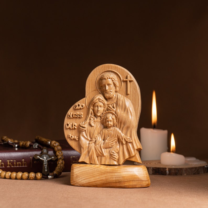 Op de afbeelding: Een houten beeldhouwwerk van de Heilige Familie, Jezus, Maria en Jozef, in een hartvormig frame. Het beeldhouwwerk staat op een houten voet en de woorden "God zegen ons huis" zijn in het frame gegraveerd. Het beeldhouwwerk staat voor twee brandende kaarsen.