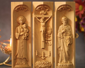 Könnte beinhalten: Ein dreiteiliges Holzrelief mit drei geschnitzten Tafeln. Die linke Tafel zeigt Unsere Liebe Frau von Lavang, die mittlere Tafel zeigt ein Kruzifix und die rechte Tafel zeigt den Heiligen Josef.