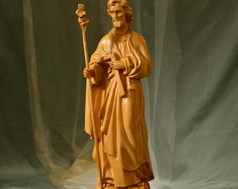 May include: A wooden statue of a saint holding a staff with a flower on top. The statue is carved in a light brown color and has a detailed face and clothing.