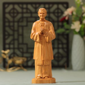 Op de afbeelding: Een houten beeld van een man in religieuze kleding, met de handen gevouwen in gebed. De figuur is gesneden uit lichtgekleurd hout, met details in de kleding en gezichtskenmerken. Het beeld staat op een rechthoekige basis.