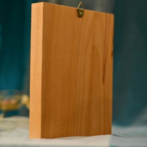 May include: A wooden frame with a light brown finish. The frame has a metal hook on the top for hanging.