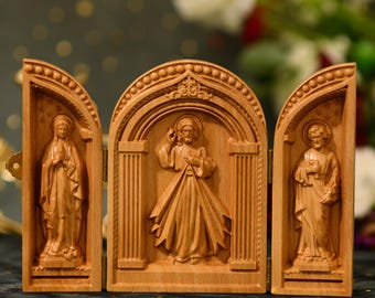 Könnte beinhalten: Ein dreiteiliges Holztriptychon mit drei geschnitzten Tafeln, die das Heilige Herz Jesu, die Jungfrau Maria und den Heiligen Johannes den Evangelisten darstellen. Die Tafeln sind mit Scharnieren versehen und können zusammengeklappt werden.