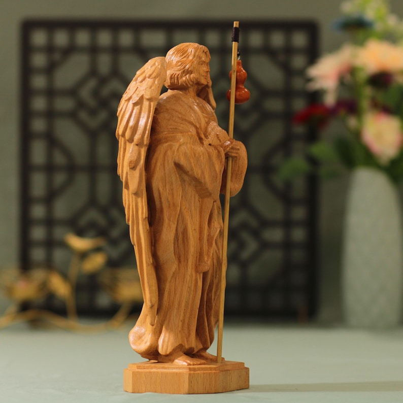 May include: A wooden sculpture of an angel with wings, holding a staff with gourds. The angel is carved in a standing position with a long robe and a staff in its right hand. The sculpture is made of light brown wood and has a natural finish.