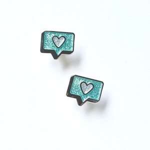 May include: Two teal blue enamel pins shaped like a speech bubble with a white heart inside. The pins are on a white background.