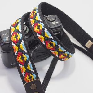 May include: A black camera strap with a colorful woven pattern. The strap has a leather adjustable piece with two brass buttons and a brass ring.