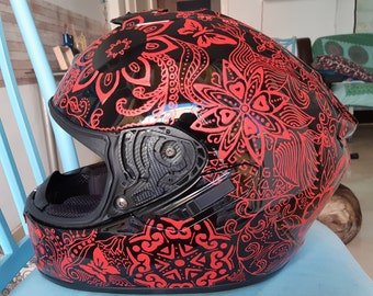 Puede incluir: Casco de moto negro brillante, adornado con intrincados patrones florales y geométricos rojos. El casco incluye una visera, ventilaciones y una correa para la barbilla. El diseño es simétrico y llamativo. Diseñado para la seguridad.