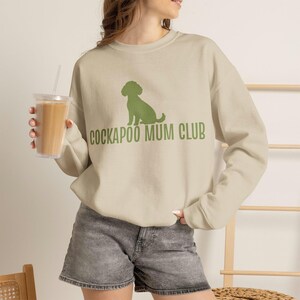 May include: A beige long-sleeve crewneck sweatshirt featuring a green graphic of a sitting dog silhouette above the text COCKAPOO MUM CLUB in a bold, sans-serif font. The garment is paired with gray denim shorts, creating a casual look.